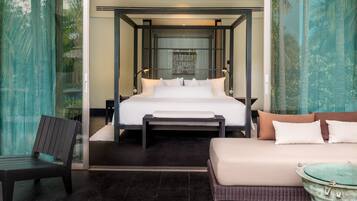 A modern hotel room with a four-poster bed, a sitting area with a bench and a small table, and a view of greenery through the window.