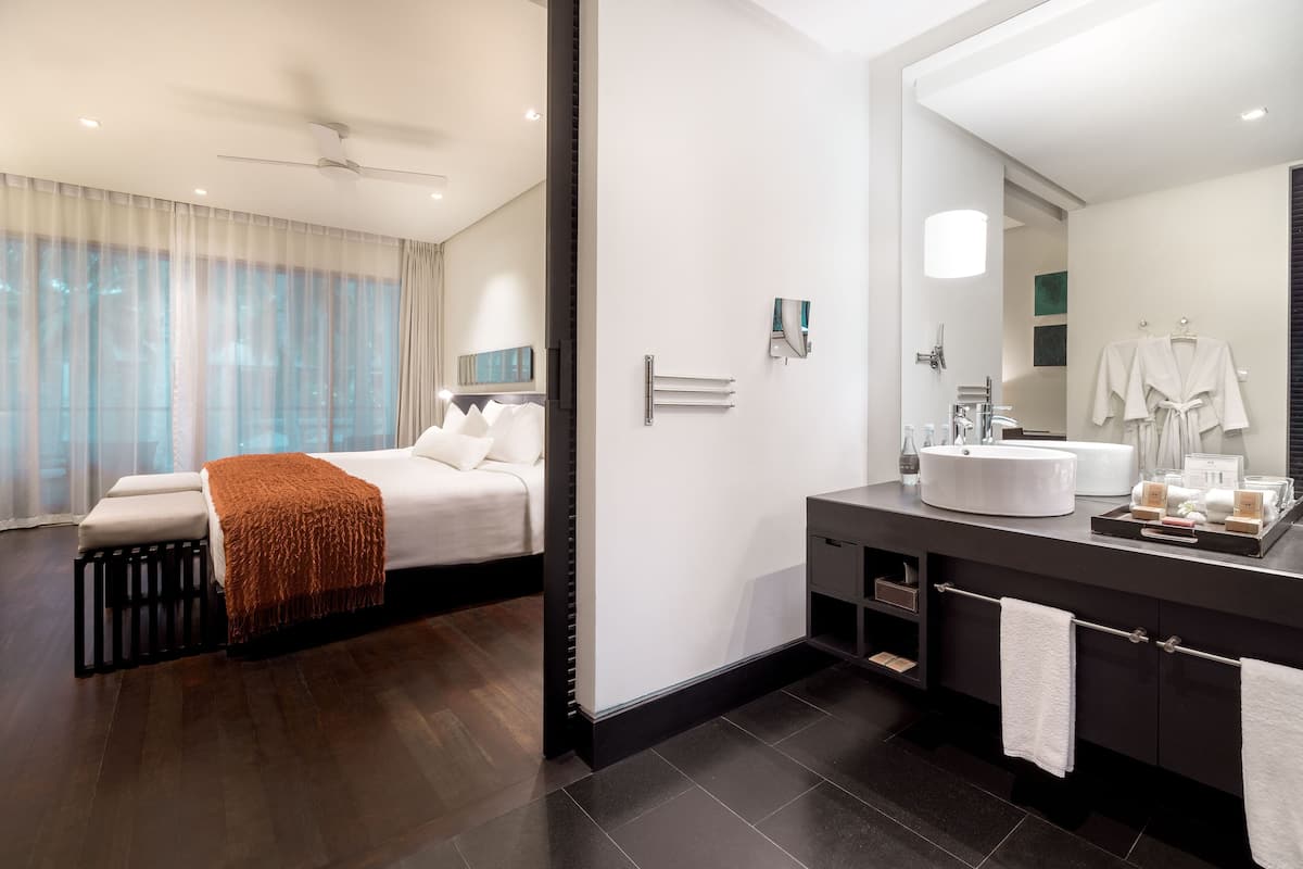 Une chambre d’hôtel moderne, dotée d’un lit, d’un grand miroir, d’une salle de bain avec un lavabo et un porte-serviettes, et offrant une vue sur l’extérieur à travers des rideaux transparents.