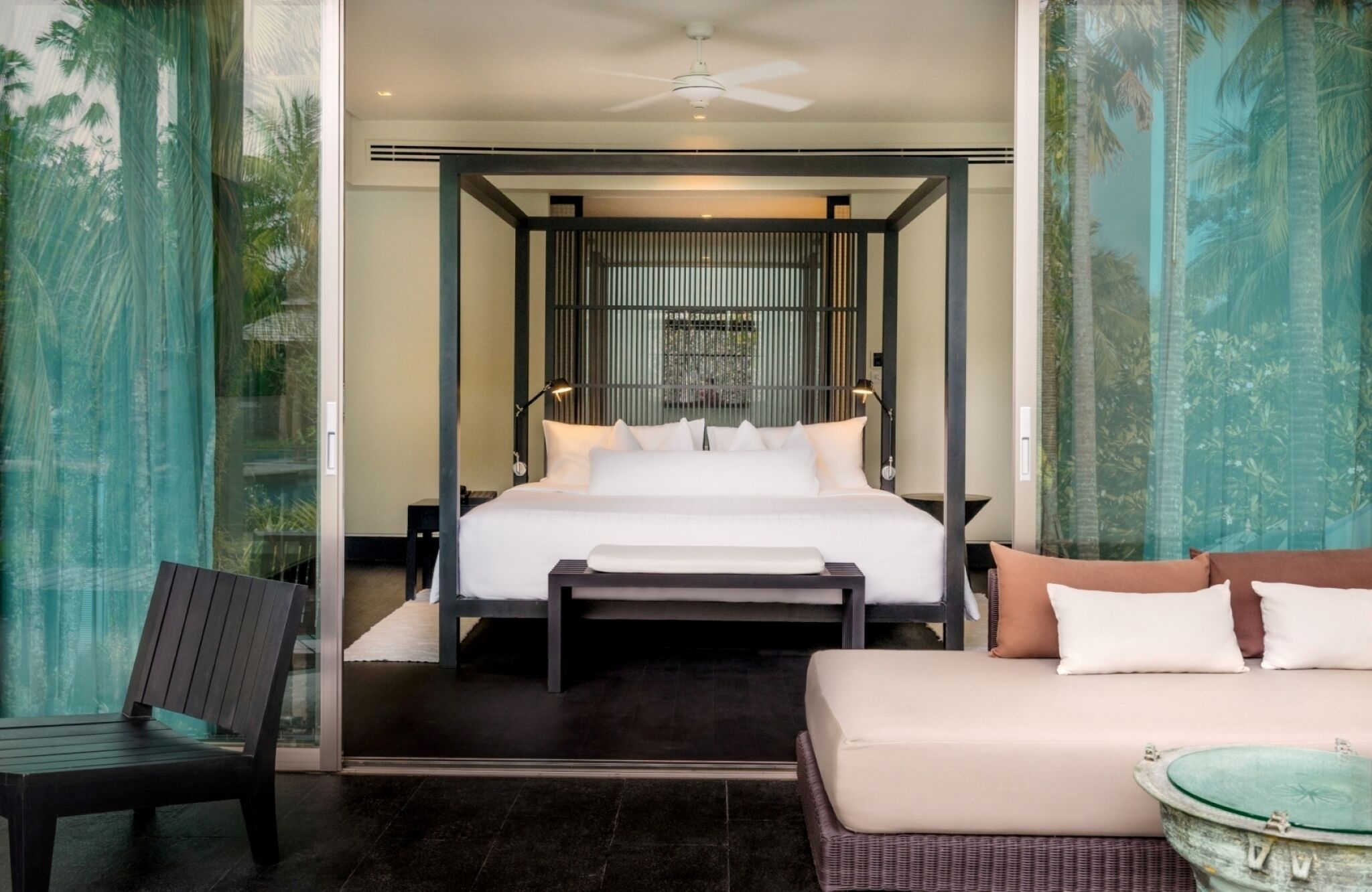 Habitación de hotel moderna con una cama grande, un sofá, una silla y vista a palmeras a través del ventanal.