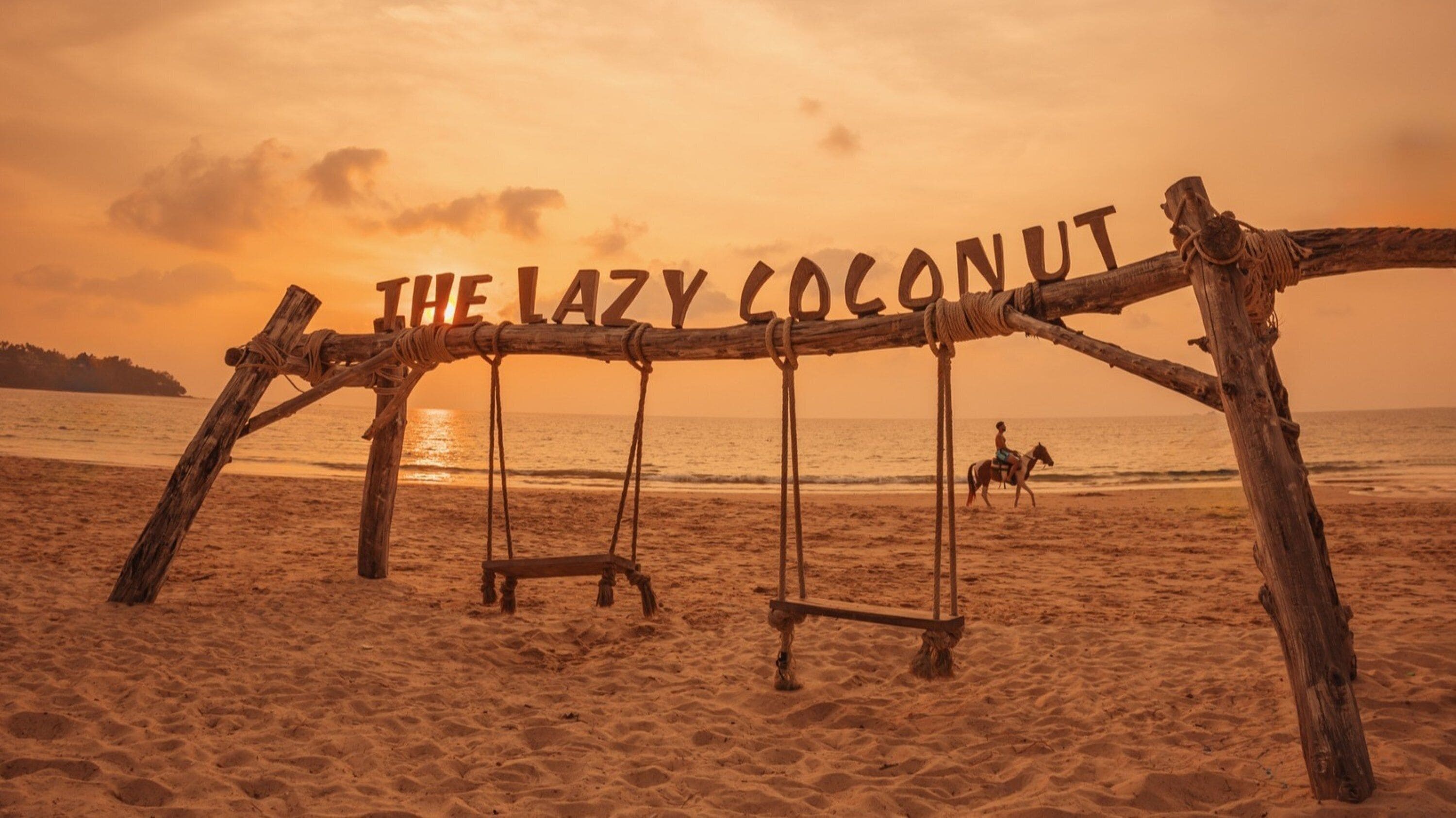 A wooden swing set with the words "THE LAZY COCONUT" written on it, set on a sandy beach with the ocean in the background.