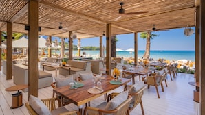 Un área de comedor al aire libre con mesas y sillas de madera, con vistas a una playa y al mar.