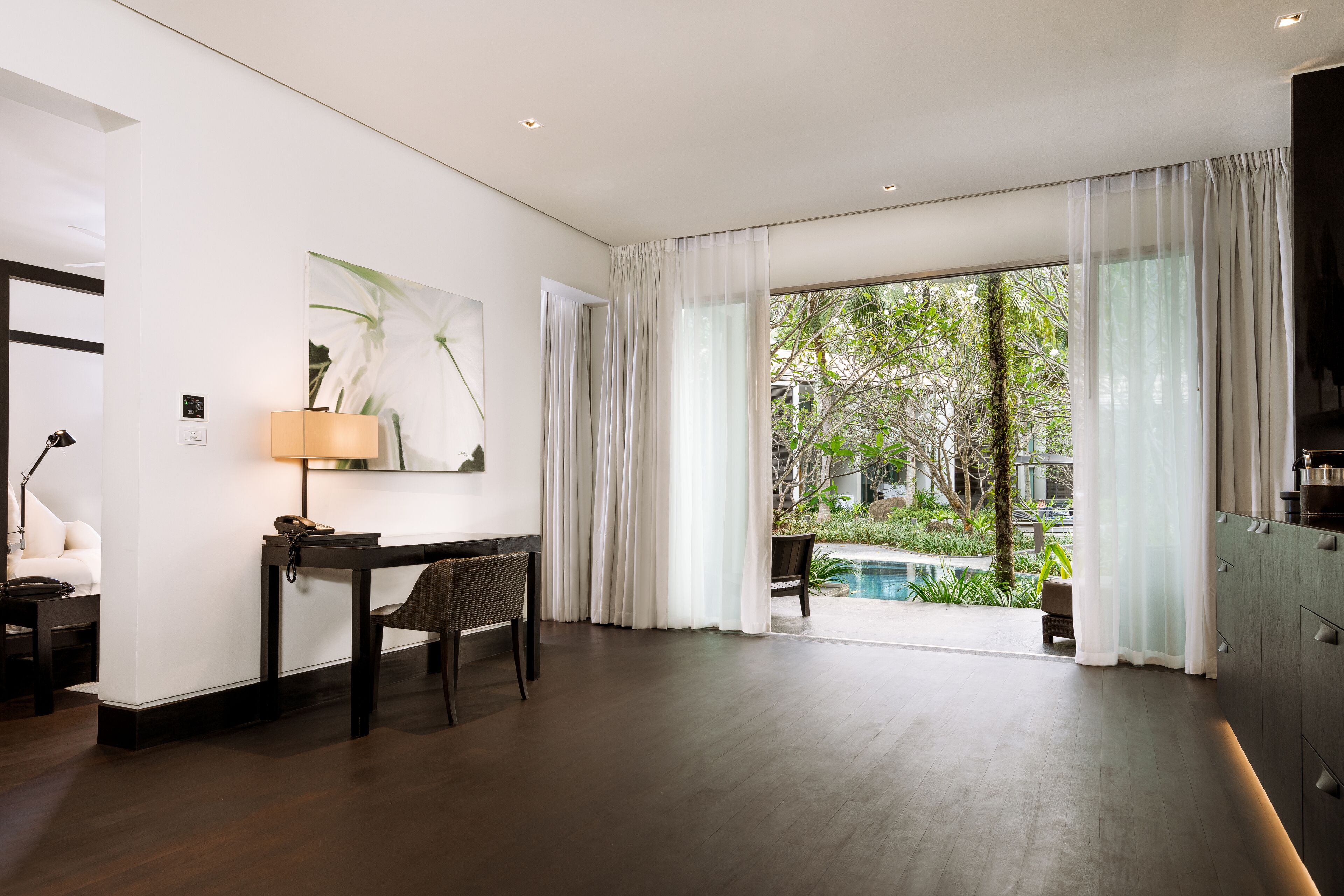 A modern hotel room with a wooden floor, a desk with a lamp, a chair, and a large window with sheer curtains.