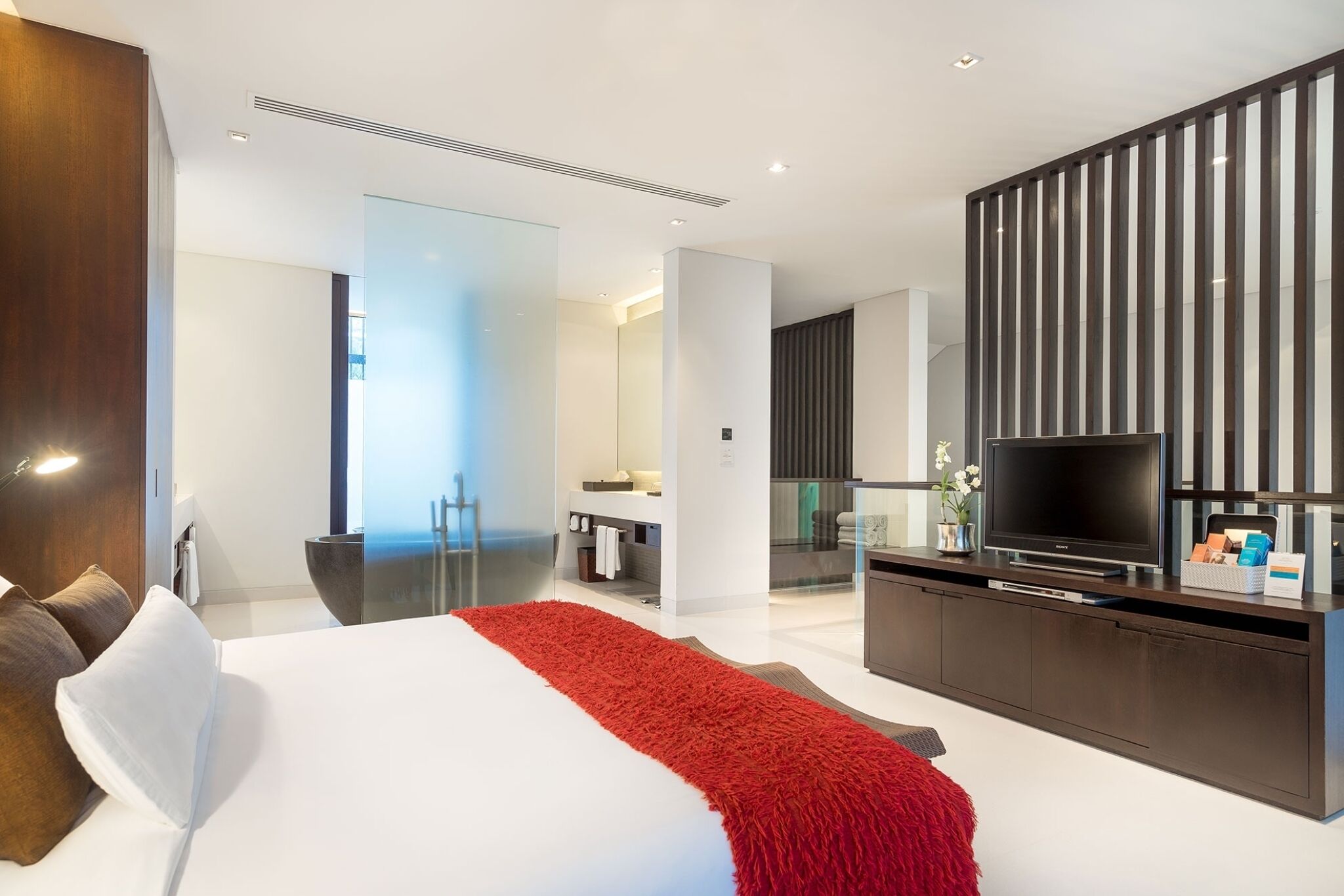 Habitación de hotel moderna con una cama grande, televisión, escritorio y baño visible a través de un panel de vidrio.