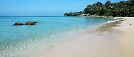 A beach with clear turquoise water, a sandy shore, and a forested hillside in the background.
