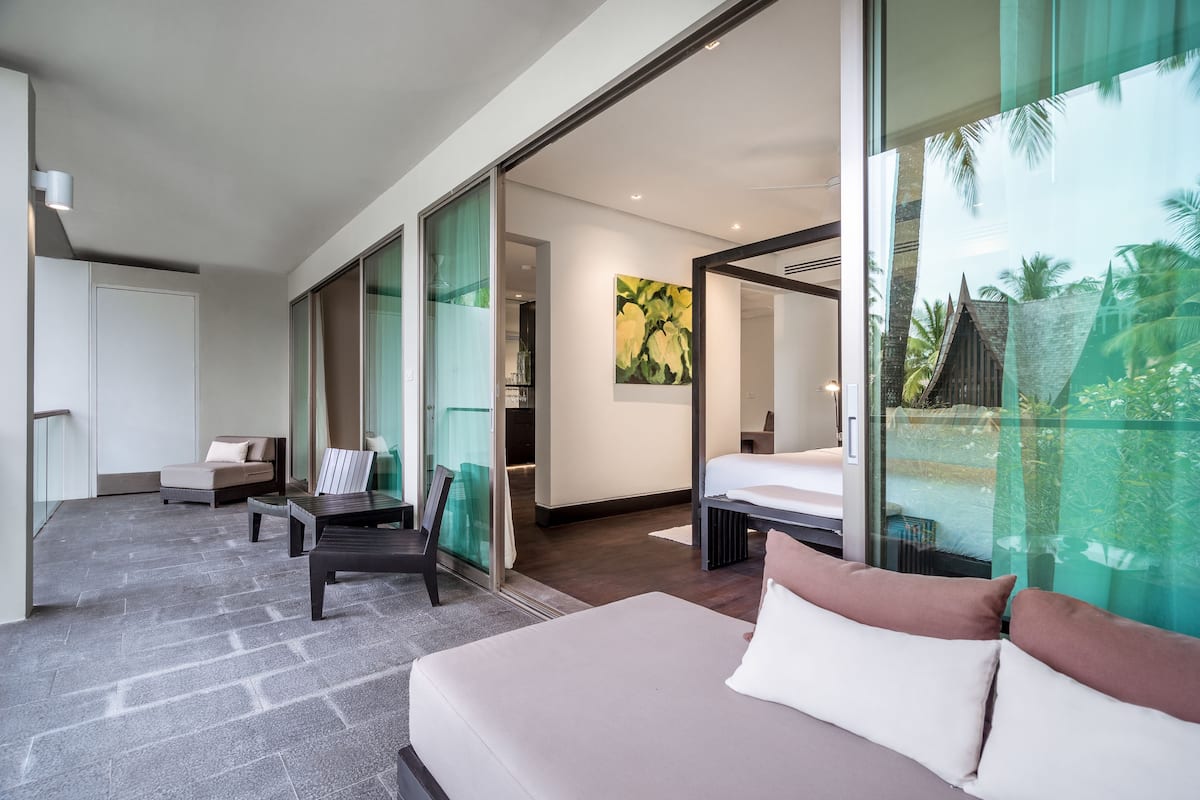 Une chambre d’hôtel moderne dotée d’un grand lit, d’un canapé, d’un bureau et d’un mur en verre offrant une vue sur les palmiers.