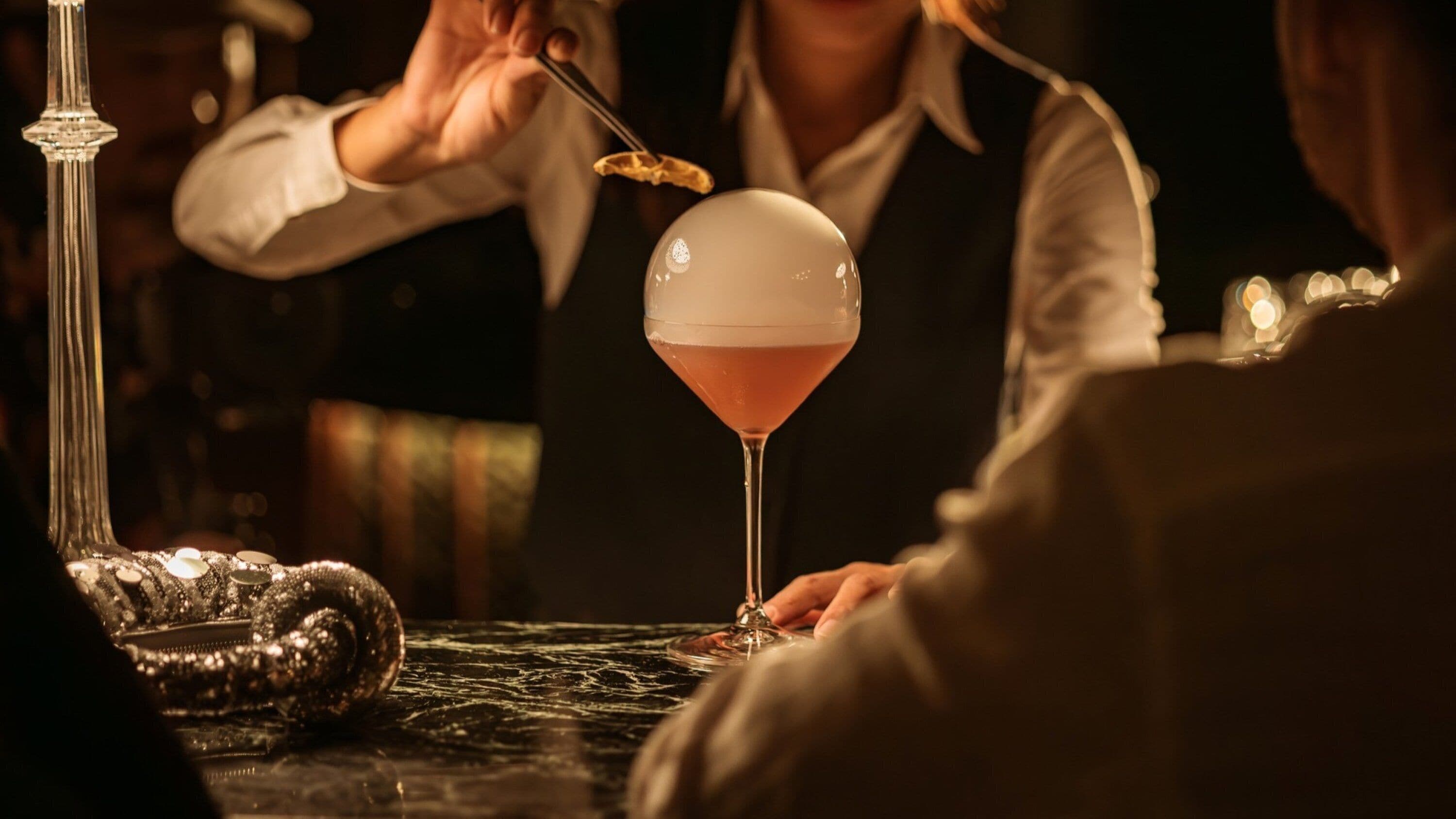 Un bar con una copa de martini sobre una mesa de mármol, una pieza decorativa de metal y una persona sosteniendo una cuchara.