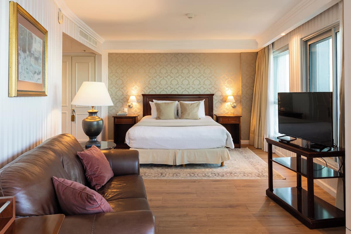 A hotel room with a bed, a leather sofa, a TV on a stand, and two bedside lamps.