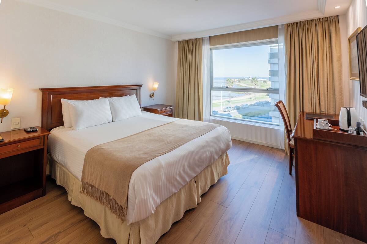 A hotel room with a large bed, wooden headboard, bedside tables with lamps, a desk with a chair, and a large window with curtains.