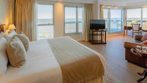 Una habitación de hotel amplia con una cama grande, televisión, zona de estar con mesa y balcón con vistas al mar.