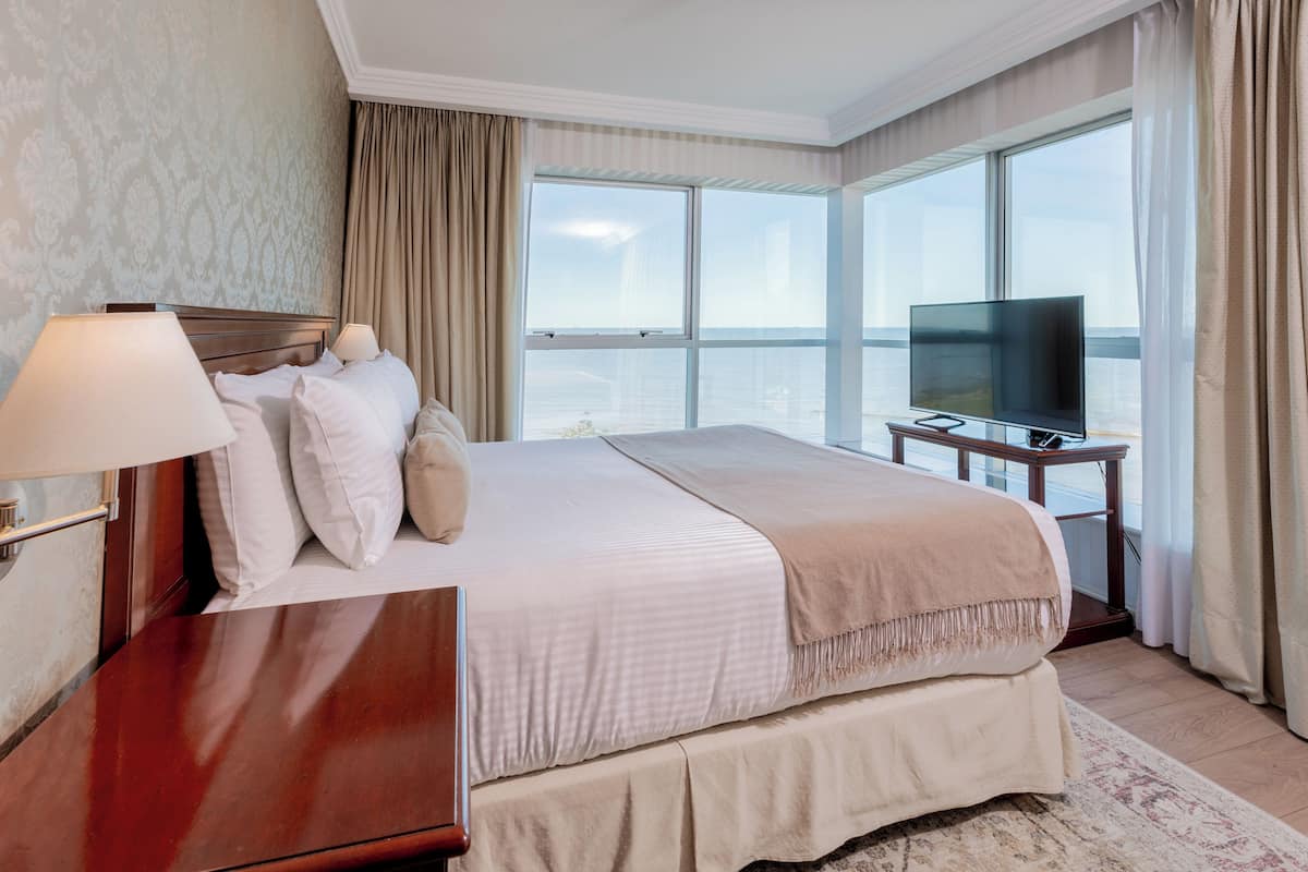A hotel room with a large bed, a TV, and a view of the city through large windows.
