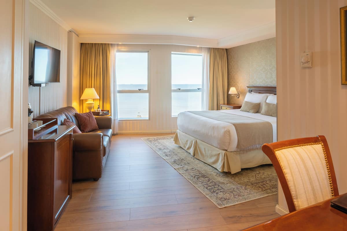 A hotel room with a bed, a sofa, a TV, a desk, and a view of the sea.