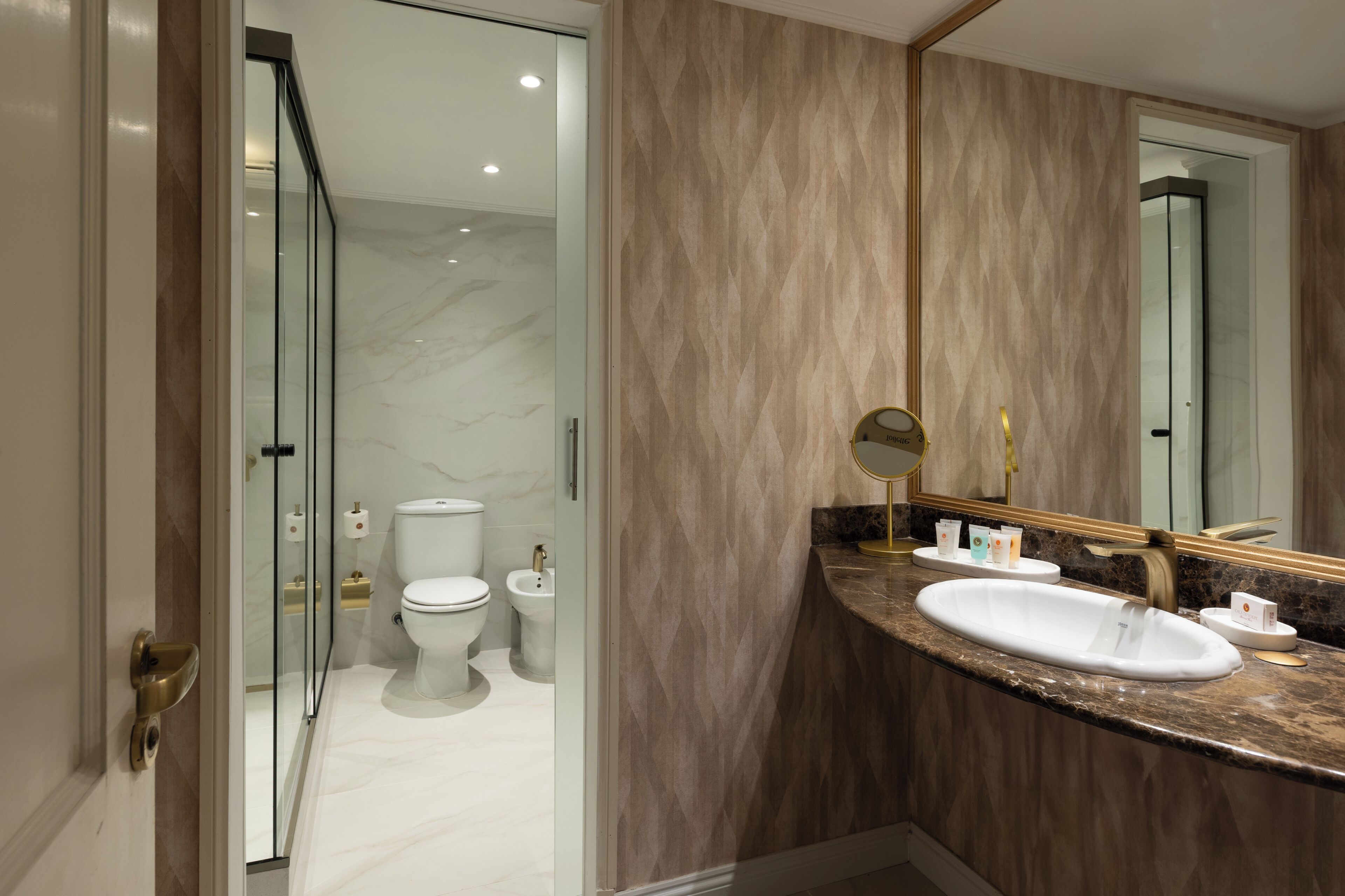 A modern bathroom with a marble floor, a white sink, a large mirror, and a glass shower enclosure.
