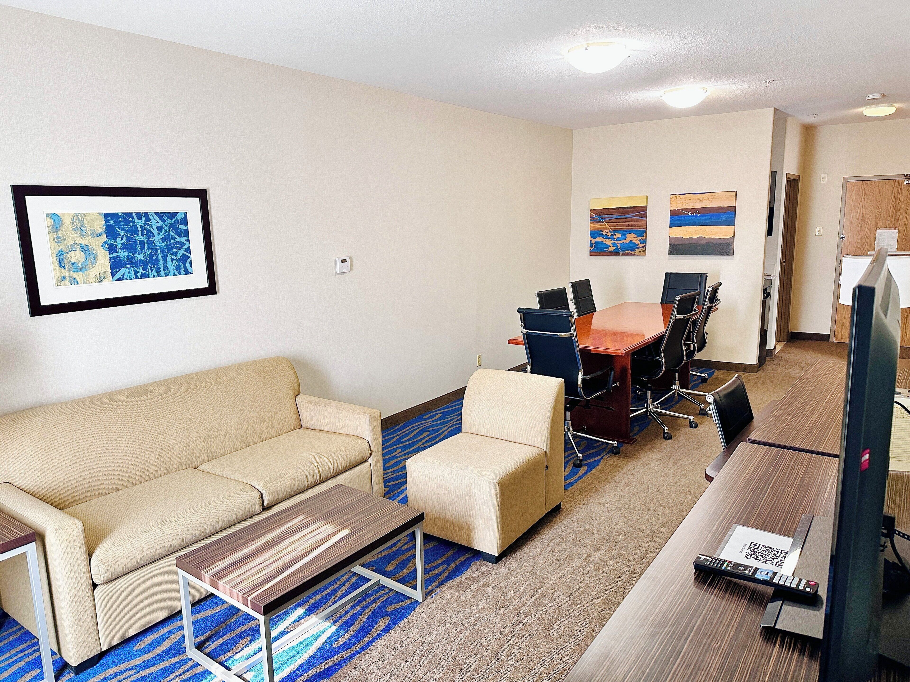 Habitación de hotel con sofá, una mesita, una mesa de conferencias con sillas, televisor y obras de arte en las paredes.