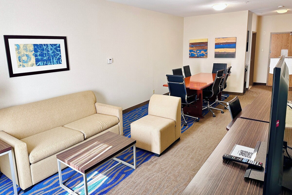 A hotel room with a sofa, a small table, a TV, a conference table with chairs, and artwork on the walls.