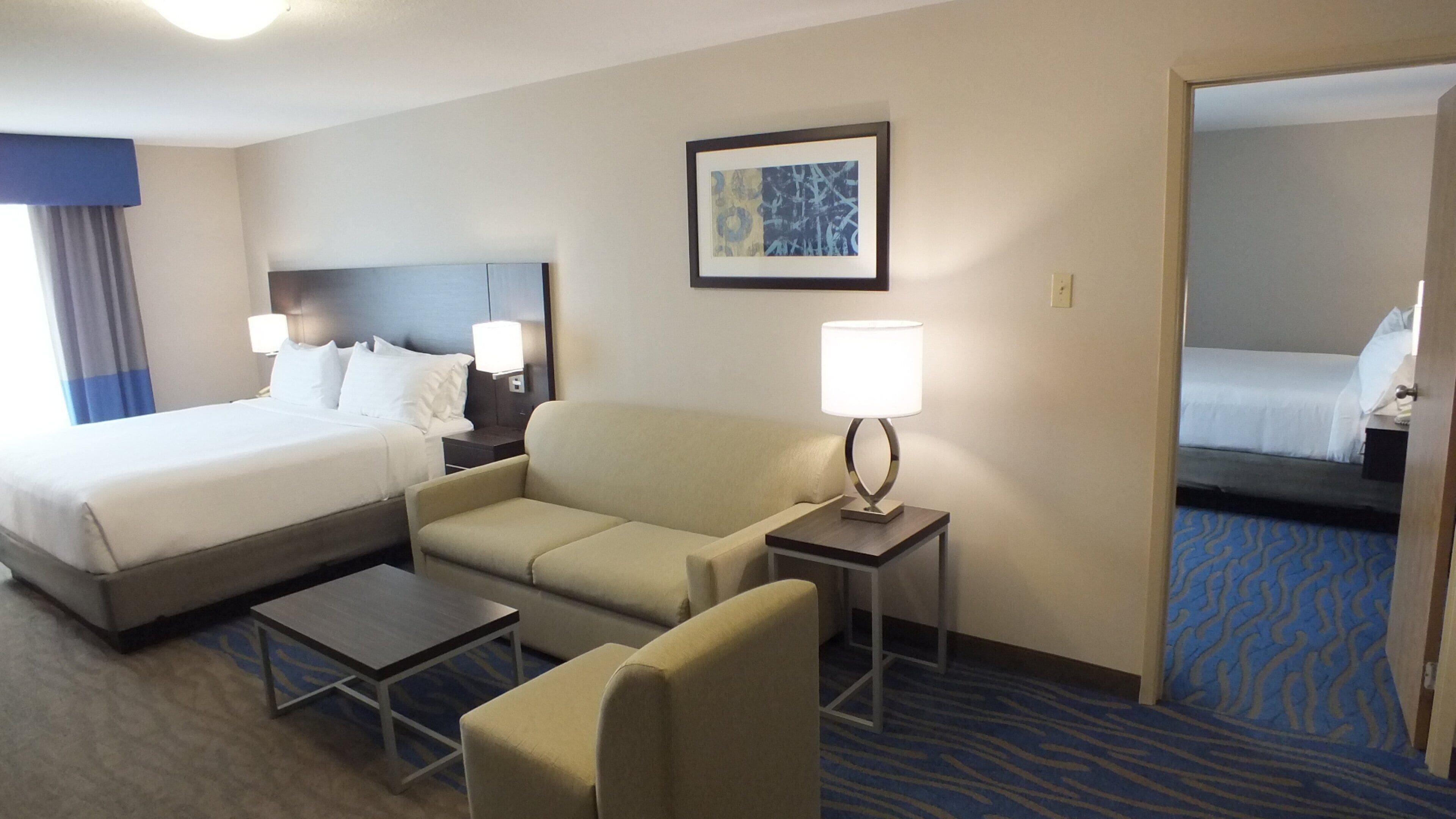 Habitación de hotel con cama, sofá, una mesita, lámpara y un cuadro en la pared.