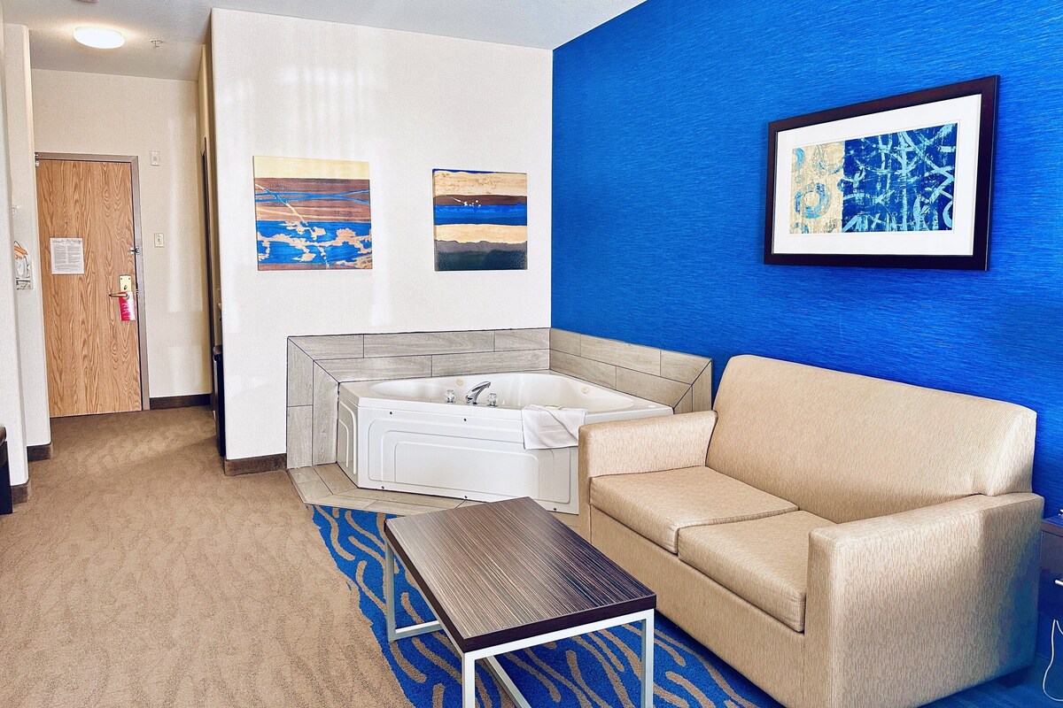 A hotel room with a beige sofa, a small table, a bathtub, and two framed artworks on the wall.
