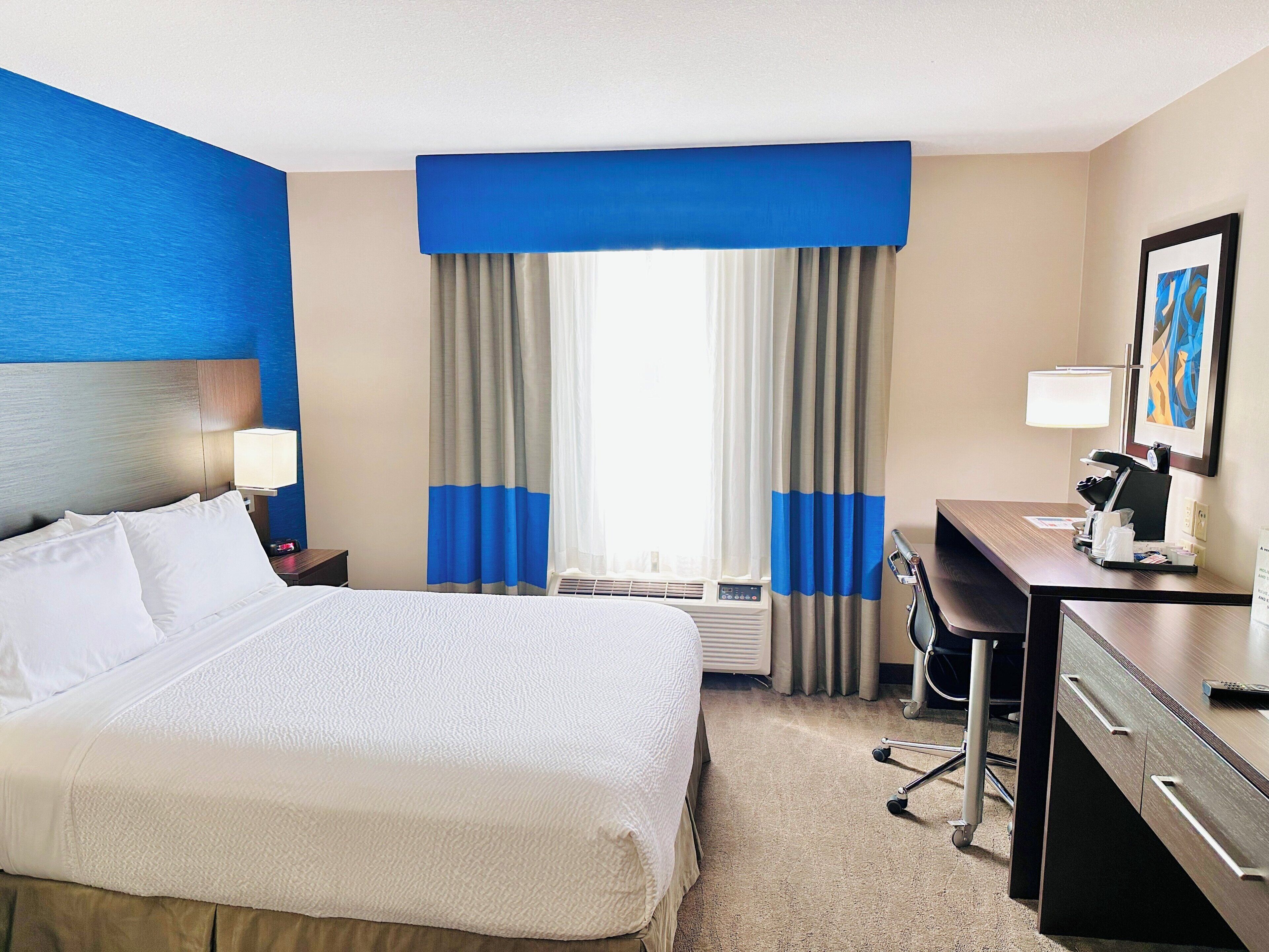 A hotel room with a large bed, a desk with a computer, a chair, a lamp, and a painting on the wall.