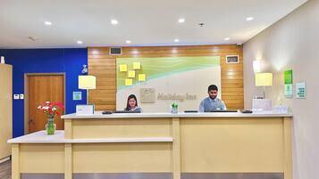 A hotel reception desk with two staff members, a wooden panel wall, and a check-in sign.