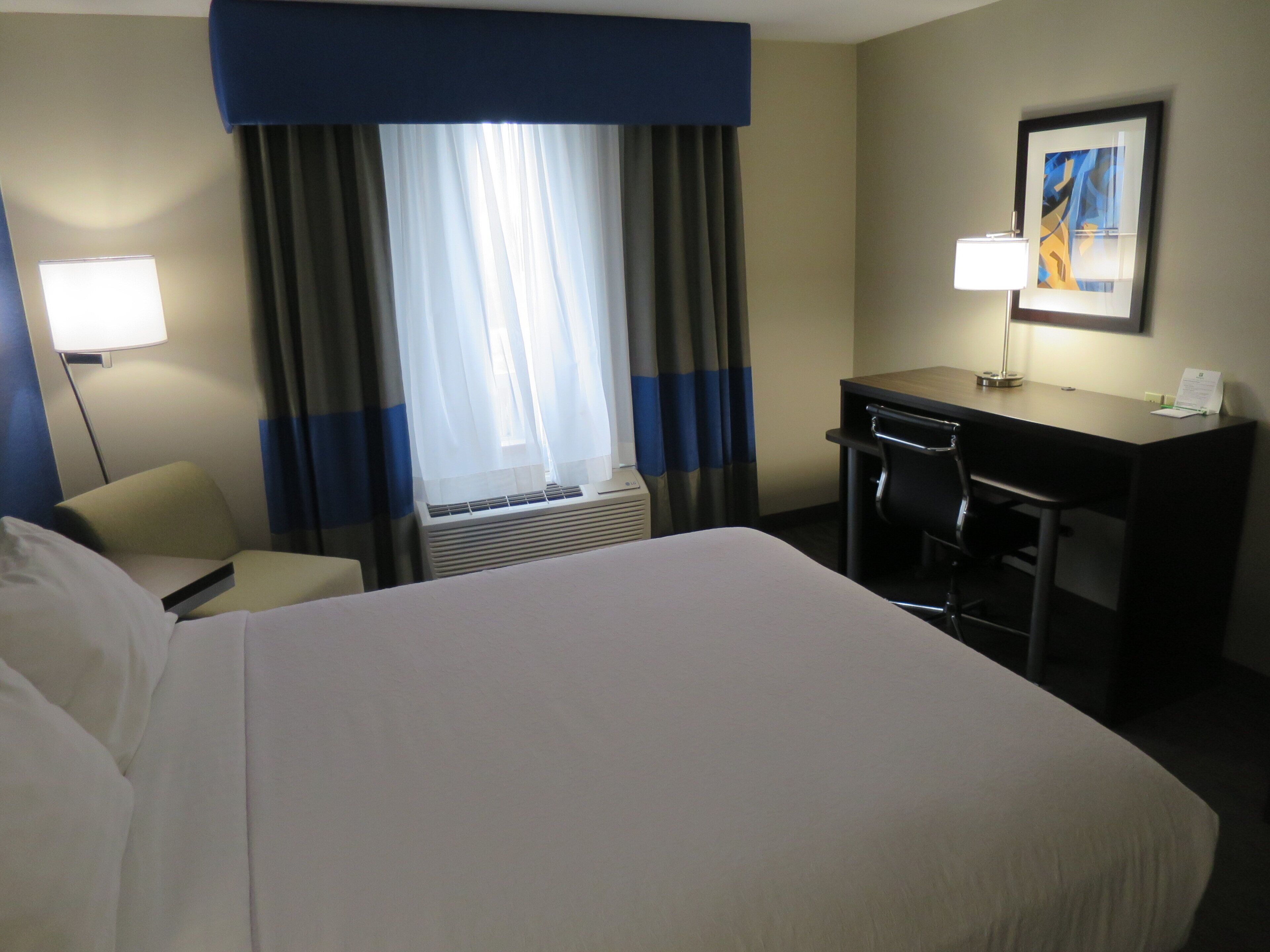A hotel room with a bed, a desk, a chair, a lamp, and a window with curtains.