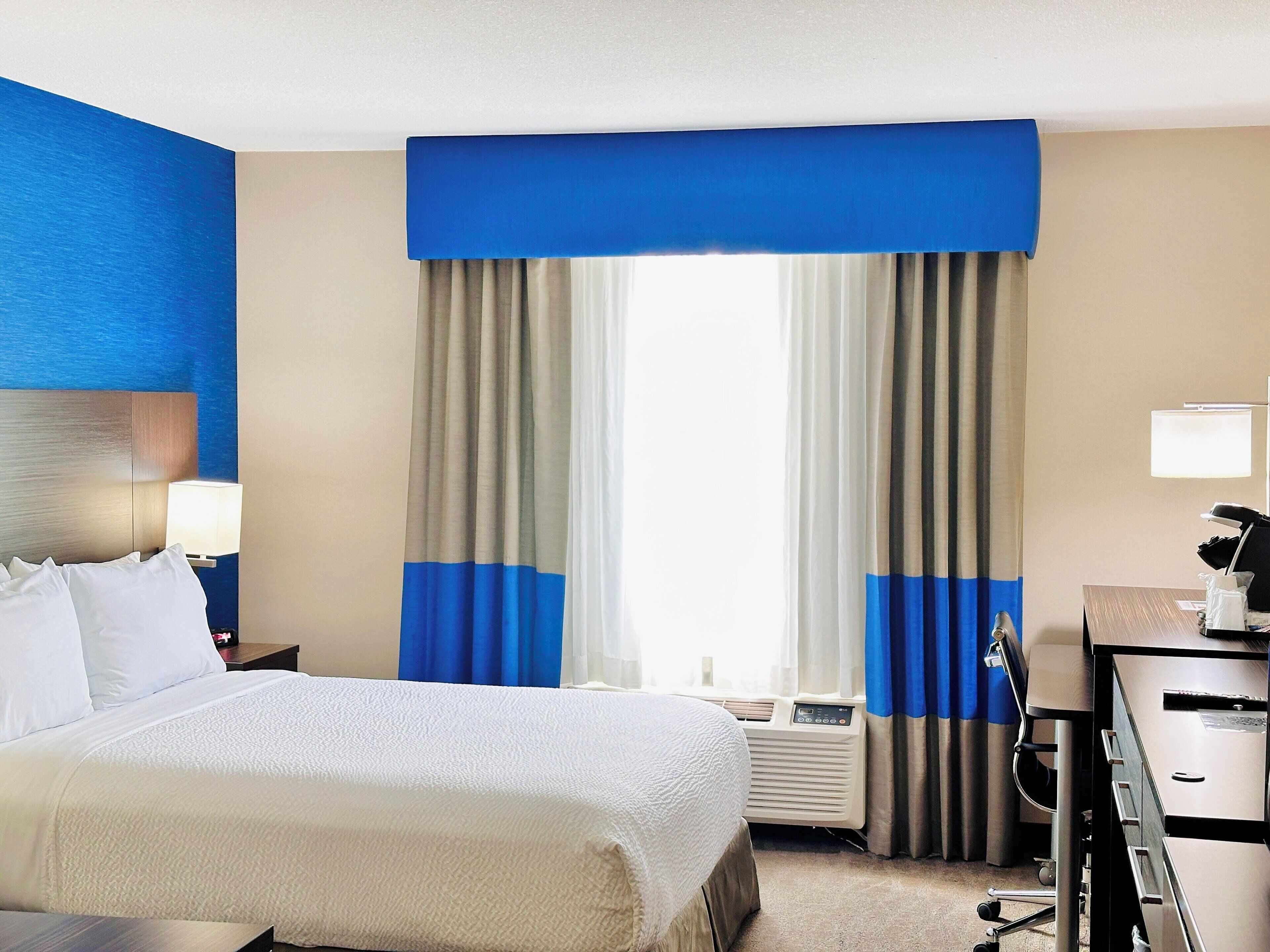 Quarto de hotel com cama, escrivaninha, cadeira, abajur e janela com cortinas.