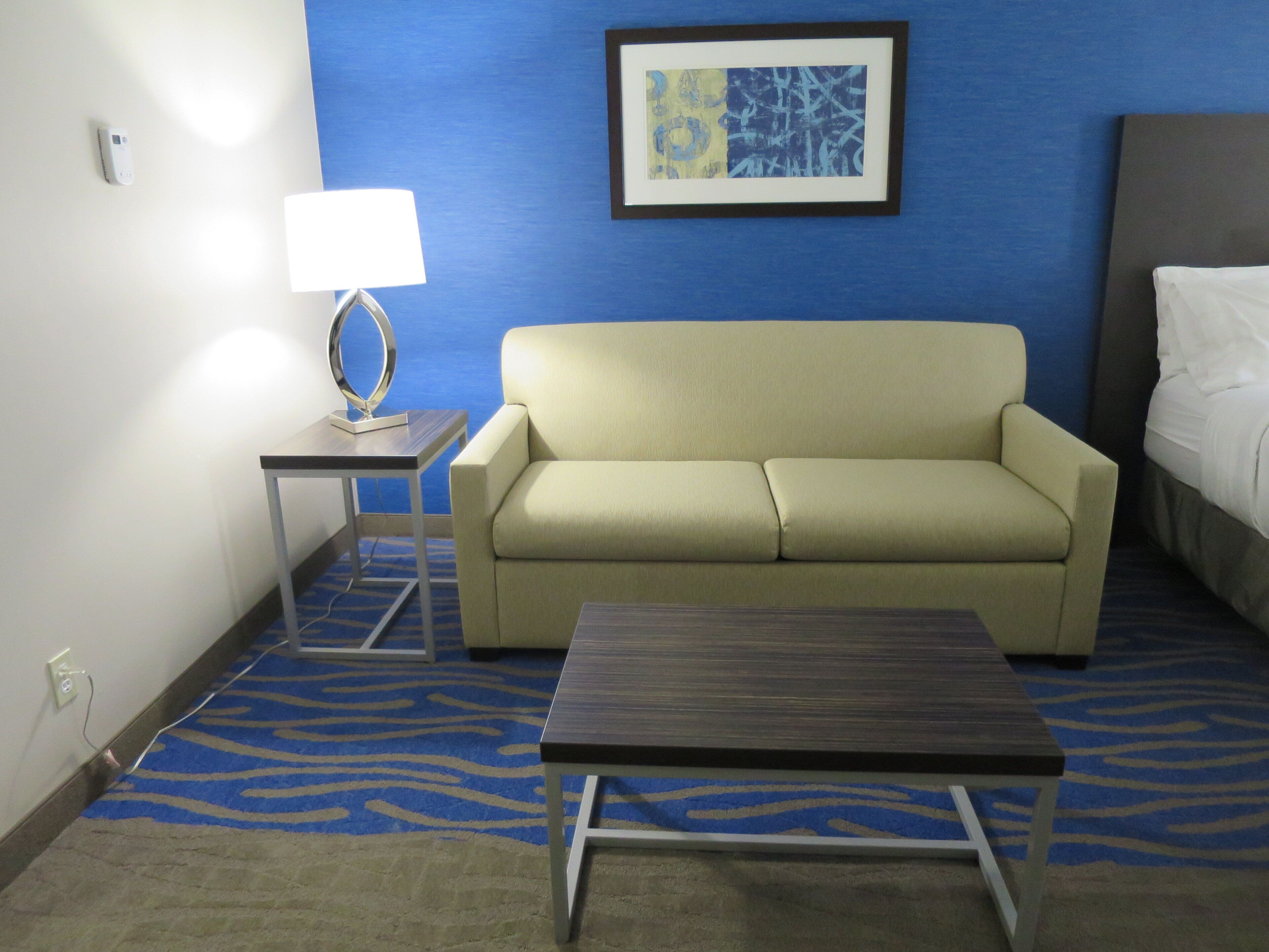 A hotel room with a sofa, a small table, a lamp, a bed, and a framed abstract painting on the wall.