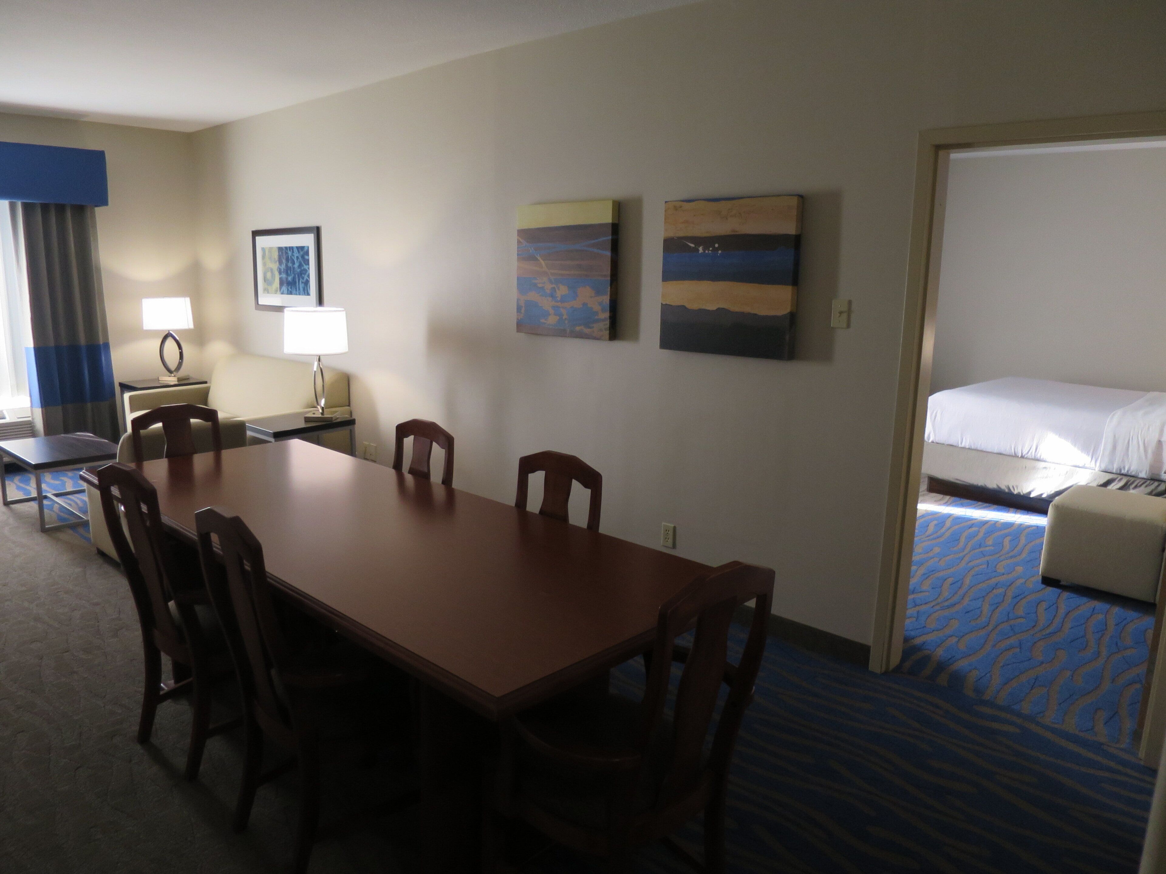 Une chambre d’hôtel avec une grande table en bois, quatre chaises, un lit, un bureau avec une lampe et deux tableaux accrochés au mur.