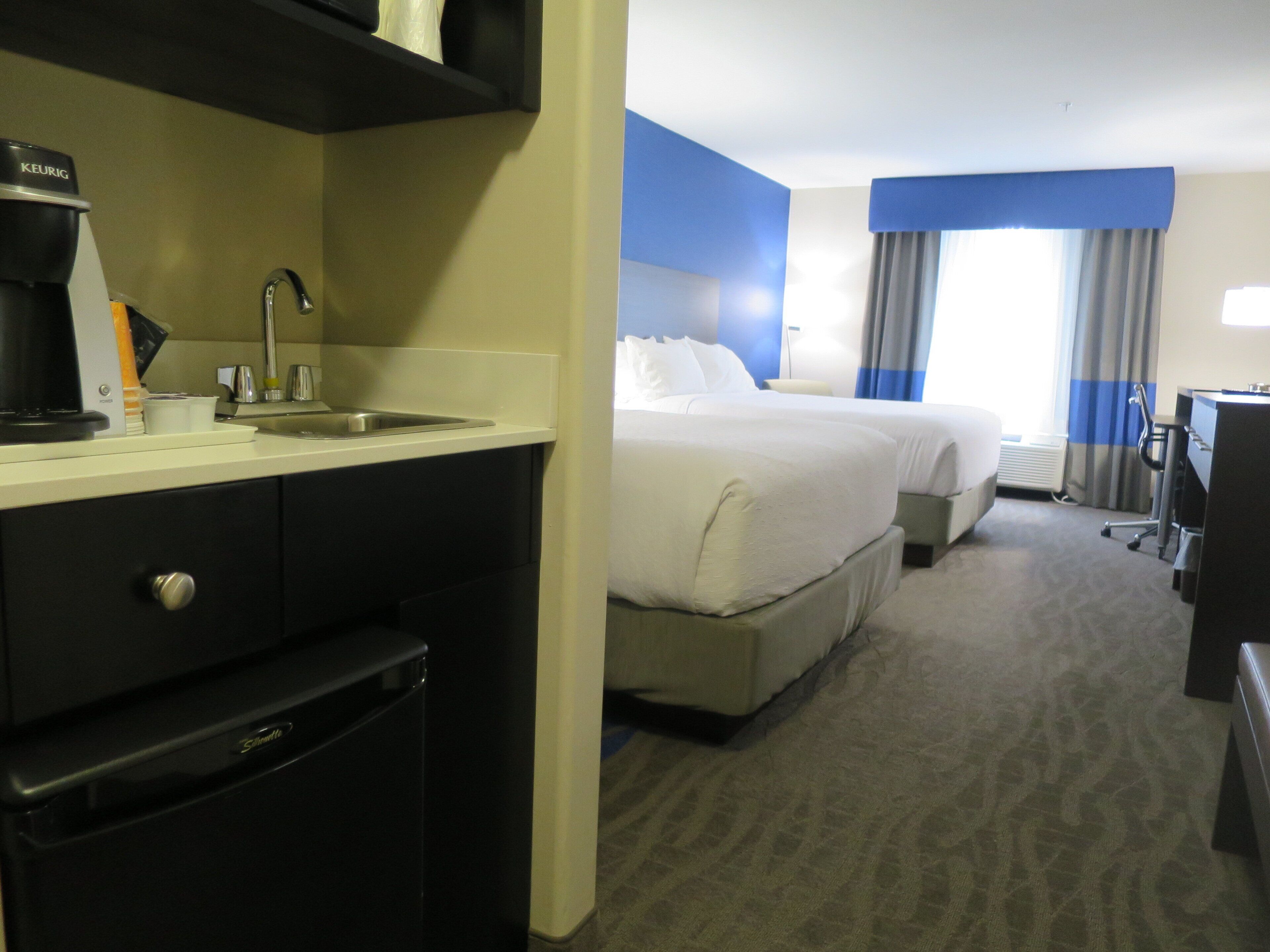 A hotel room with a kitchenette, a bed, a desk, and a window with curtains.