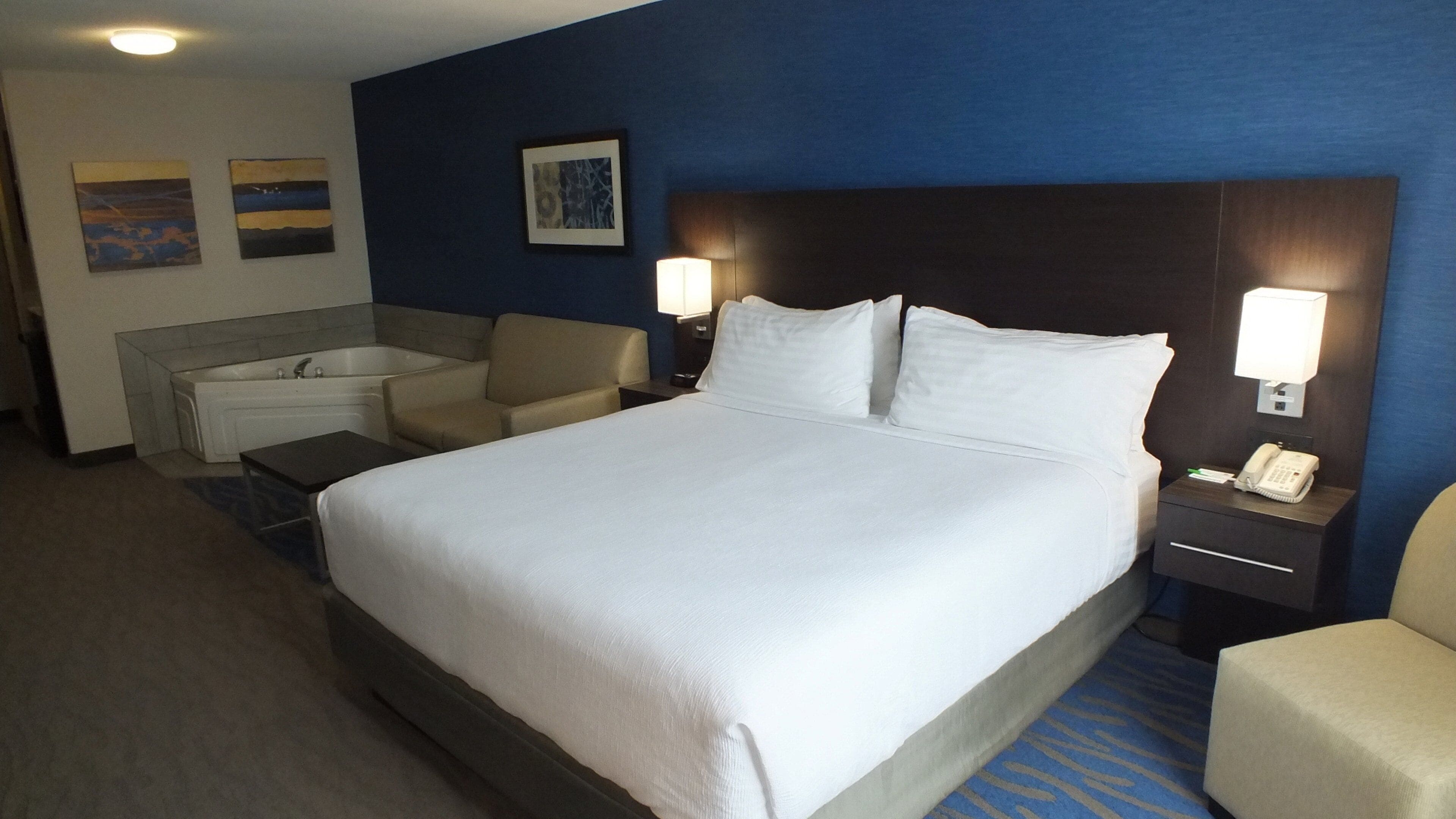 Ein Hotelzimmer mit einem großen Bett, einer Sitzecke mit Sessel und kleinem Tisch, einer Badewanne sowie einem Schreibtisch mit Telefon und Lampe.