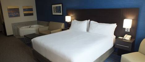 A hotel room with a large bed, a sitting area with a chair and a small table, a bathtub, and a desk with a phone and lamp.