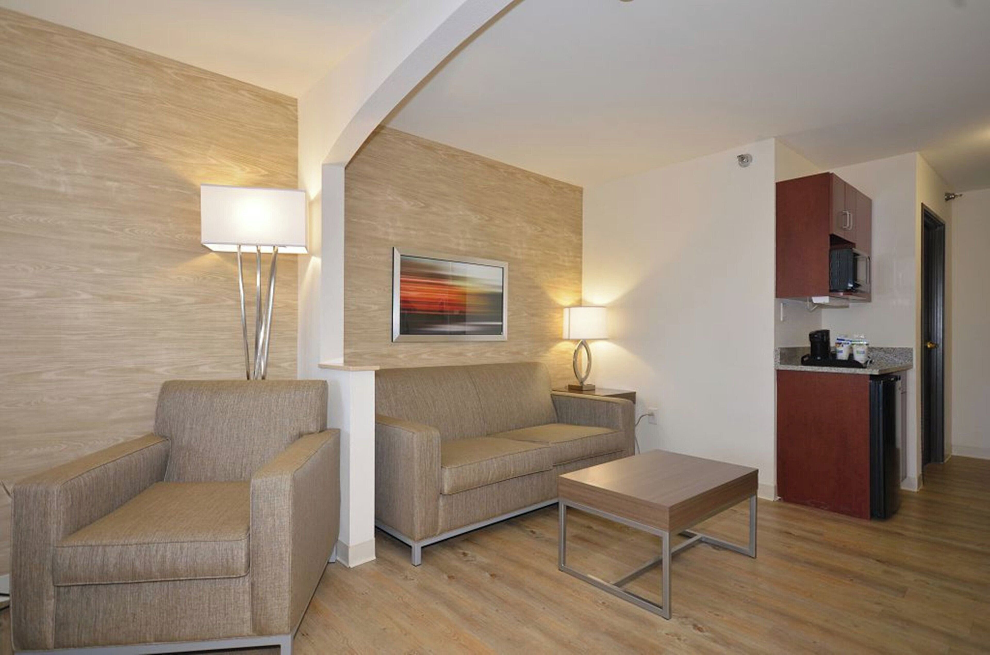Een moderne woonkamer met een bank, fauteuil en salontafel, een schilderij aan de muur, en een kitchenette op de achtergrond.