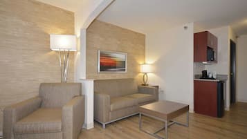 Een moderne woonkamer met een bank, fauteuil en salontafel, een schilderij aan de muur, en een kitchenette op de achtergrond.
