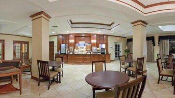 Um saguão de hotel com um balcão de recepção central, várias áreas de estar e piso ladrilhado.