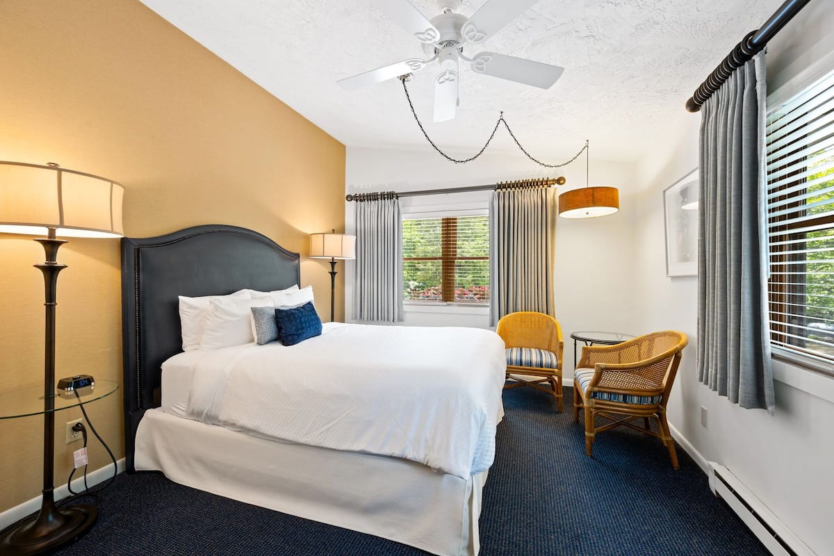 A hotel room with a bed, two lamps, a ceiling fan, a window with curtains, and a chair.