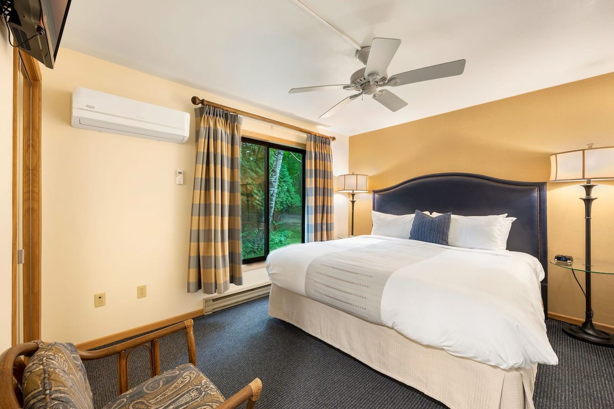 A hotel room with a bed, a ceiling fan, a window with curtains, a lamp, and a TV.