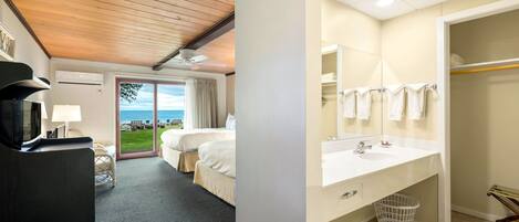 Habitación de hotel con techo de madera, cama grande, vista al mar, baño con lavamanos y espejo, y cesto de ropa.