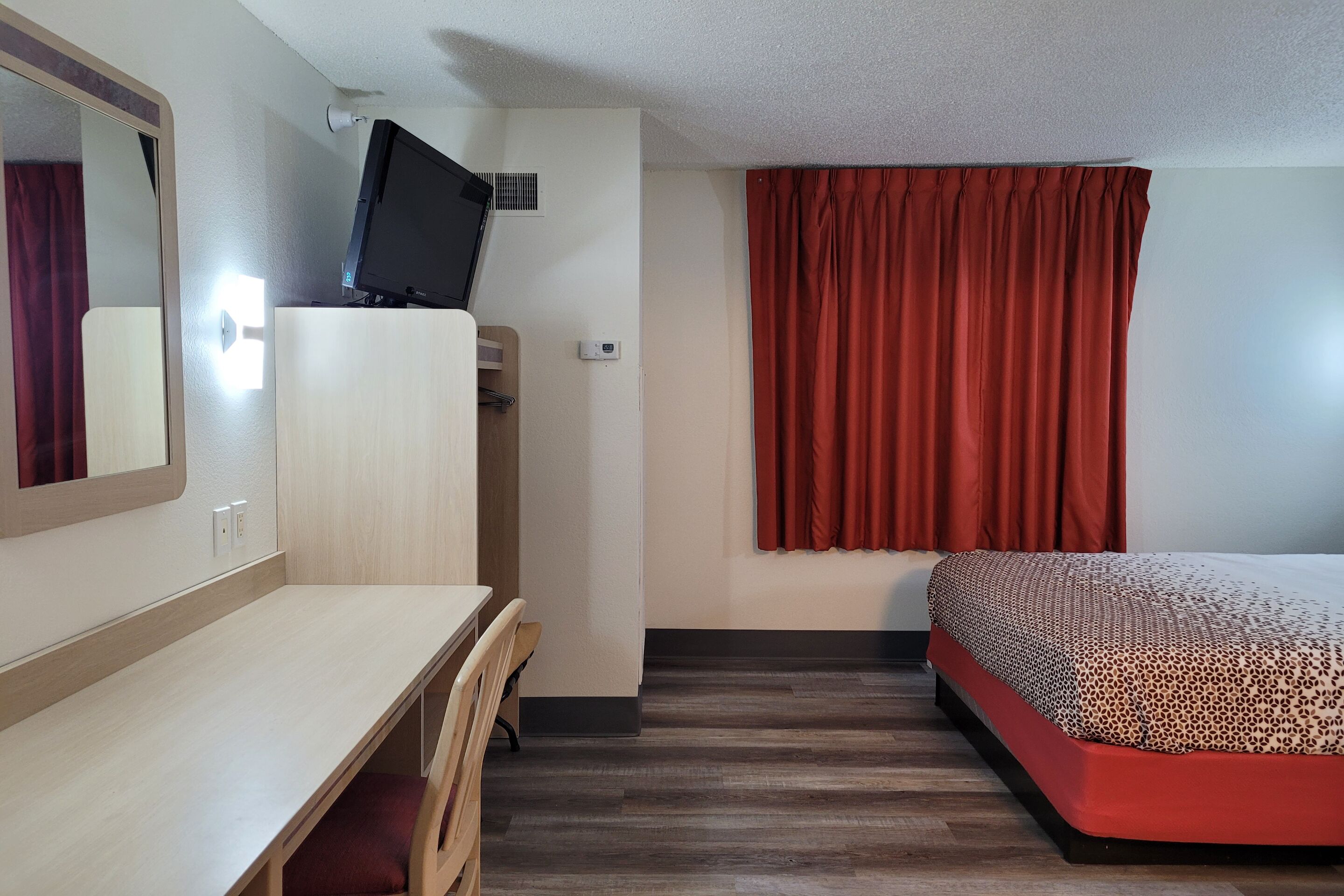 A hotel room with a wooden desk, a bed with a patterned bedspread, a television on a stand, and red curtains.