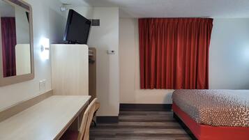 A hotel room with a wooden desk, a bed with a patterned bedspread, a television on a stand, and red curtains.