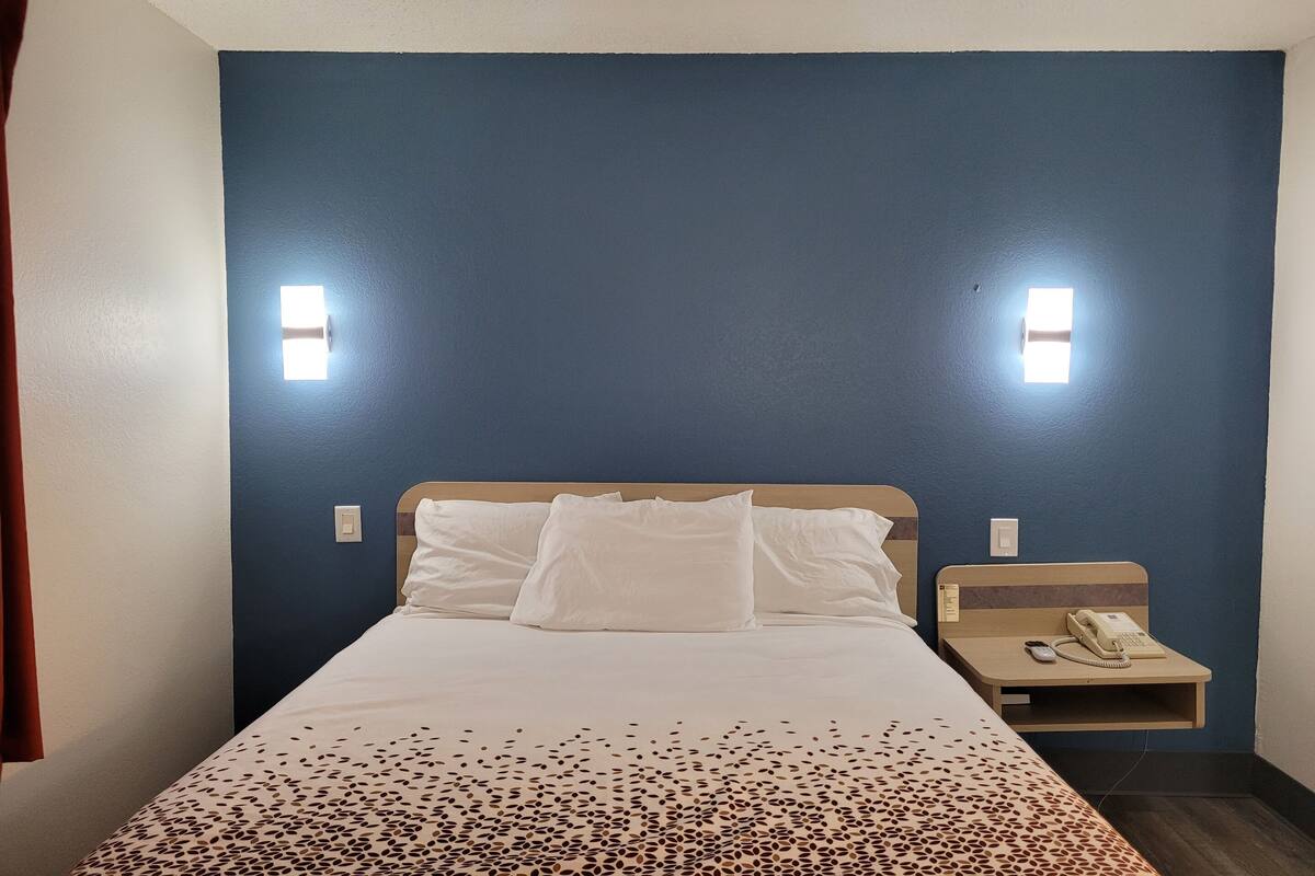 A hotel room with a bed, white bedding, a telephone on a bedside table, and two wall-mounted light fixtures.