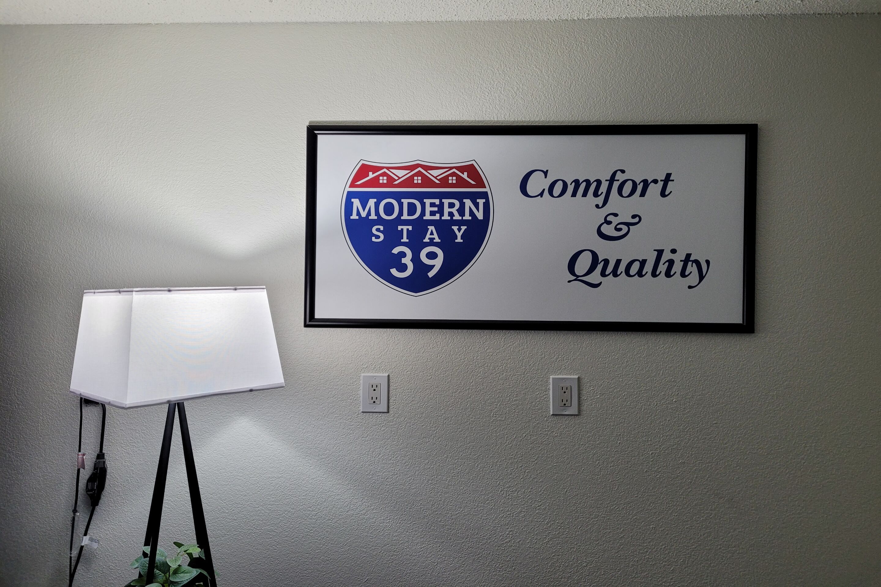 A framed sign with "Modern Stay 39" and "Comfort & Quality" text, a lamp, and two electrical outlets on a wall.