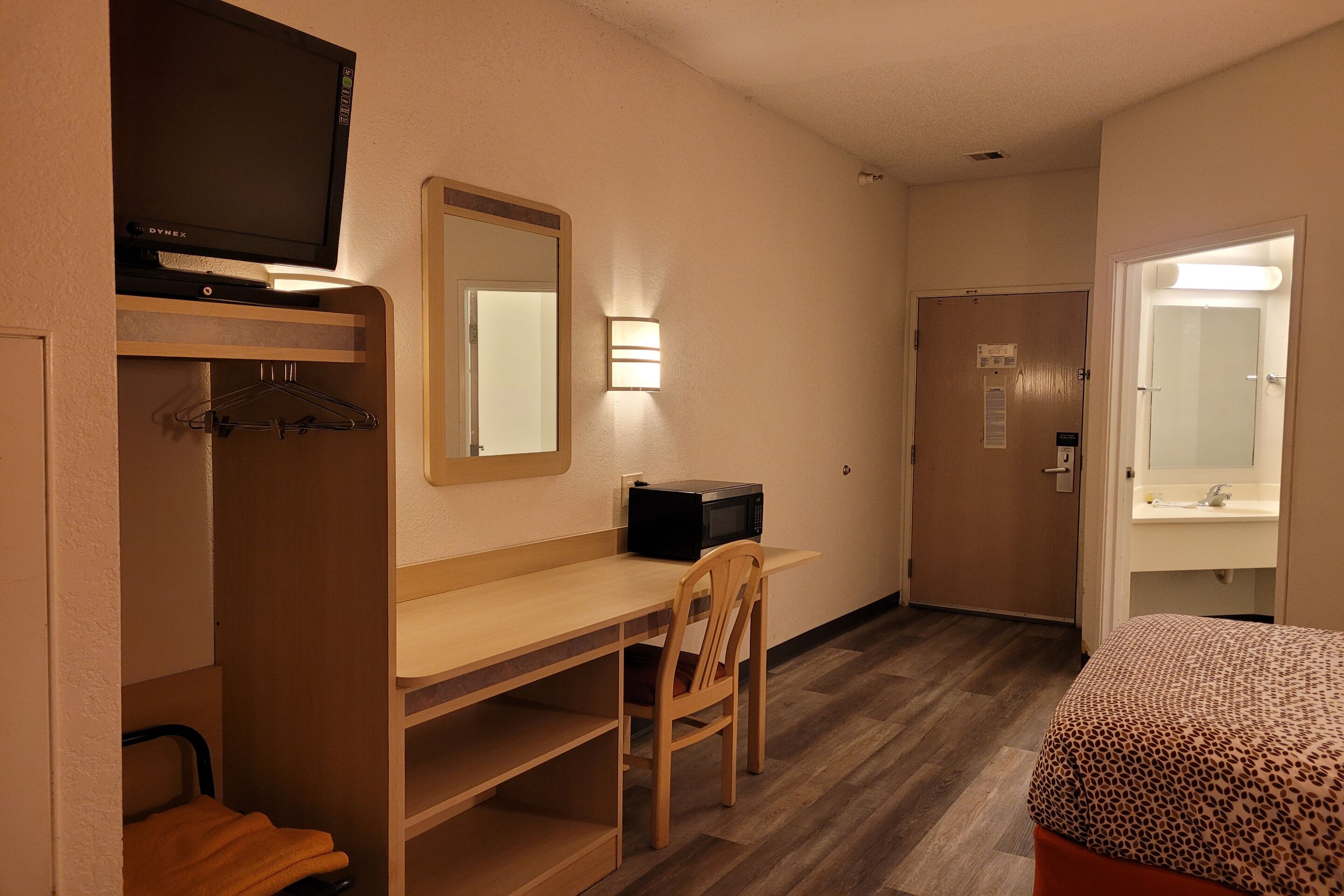 A hotel room with a wooden desk, a television, a mirror, a bed with a patterned cover, a wardrobe, and a bathroom with a mirror and sink.