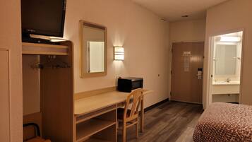 A hotel room with a wooden desk, a television, a mirror, a bed with a patterned cover, a wardrobe, and a bathroom with a mirror and sink.