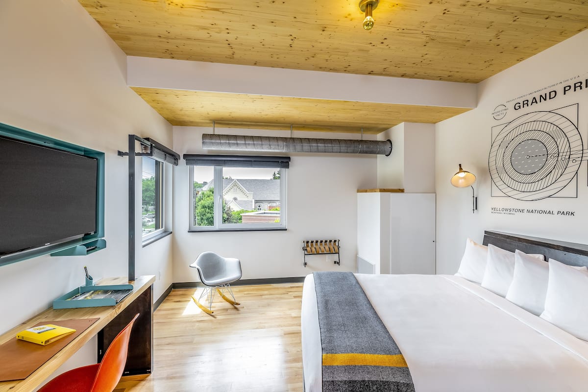 Ett hotellrum med en säng, ett skrivbord med en stol, en tv och ett fönster med utsikt över ett hus.