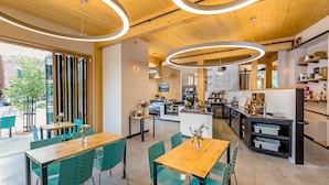 Un café moderno con mesas de madera y sillas verdes, una zona de cocina y vistas a la calle.