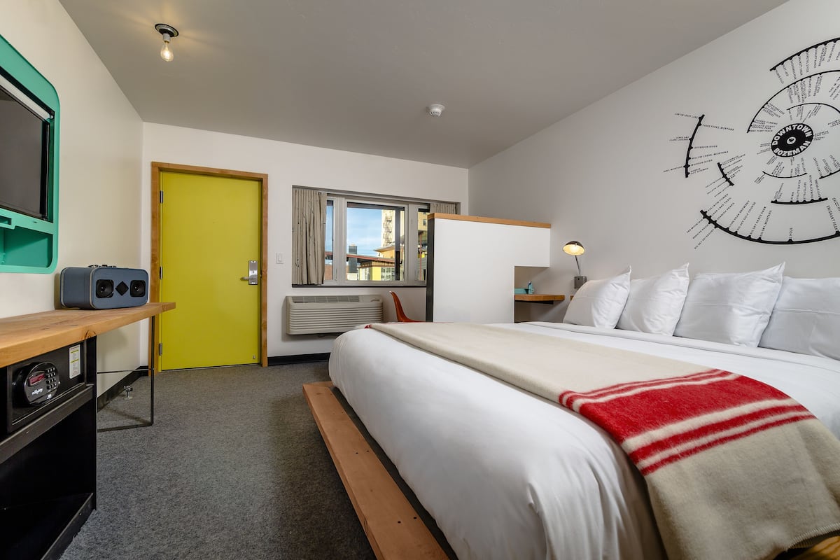 Ett hotellrum med en säng, ett skrivbord med en dator, en gul dörr och en väggklocka.
