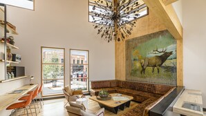 Una sala de estar moderna con techo de madera, un cuadro grande de un alce, y un comedor con sillas naranjas y una mesa de madera.