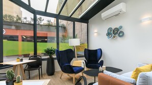 Un espacio de oficina moderno con ventanas amplias, un equipo de aire acondicionado blanco y negro, una mesa redonda, dos sillas azules, un sofá y un adorno en la pared.