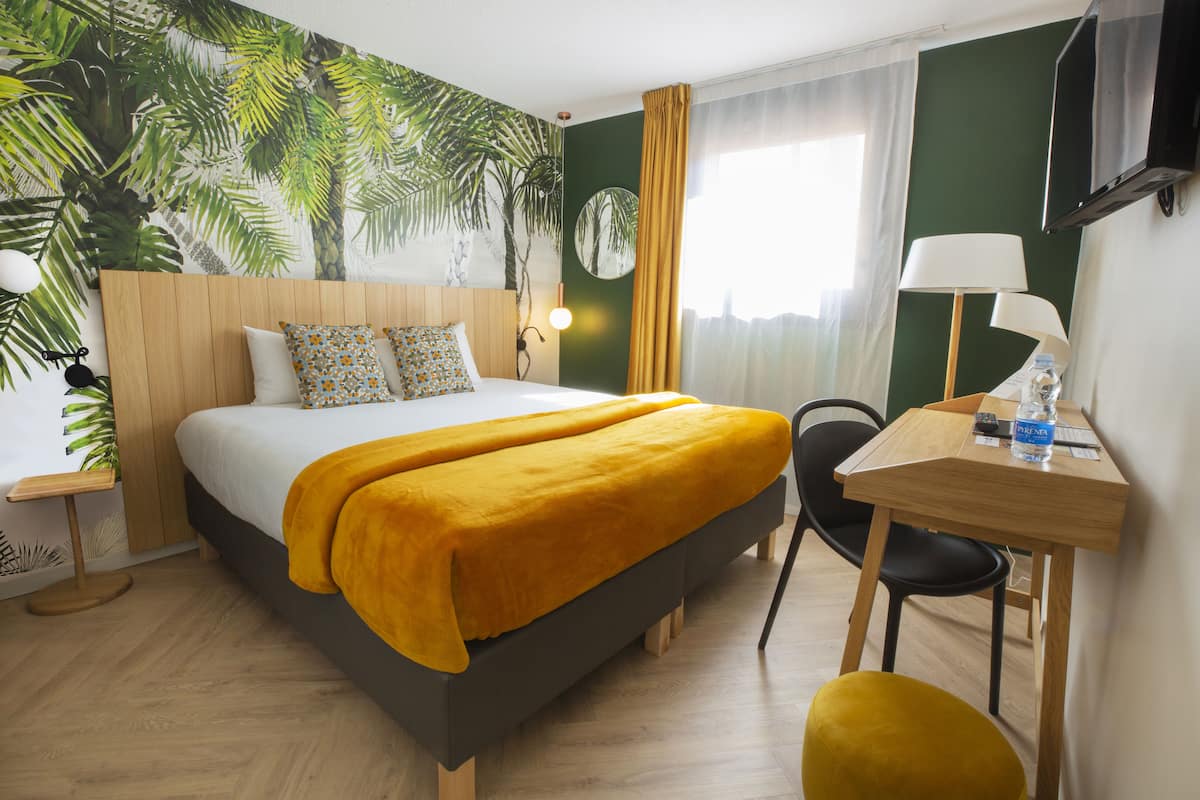 Habitación de hotel con cama, escritorio, silla, lámpara, televisor y un ventanal grande con cortinas.