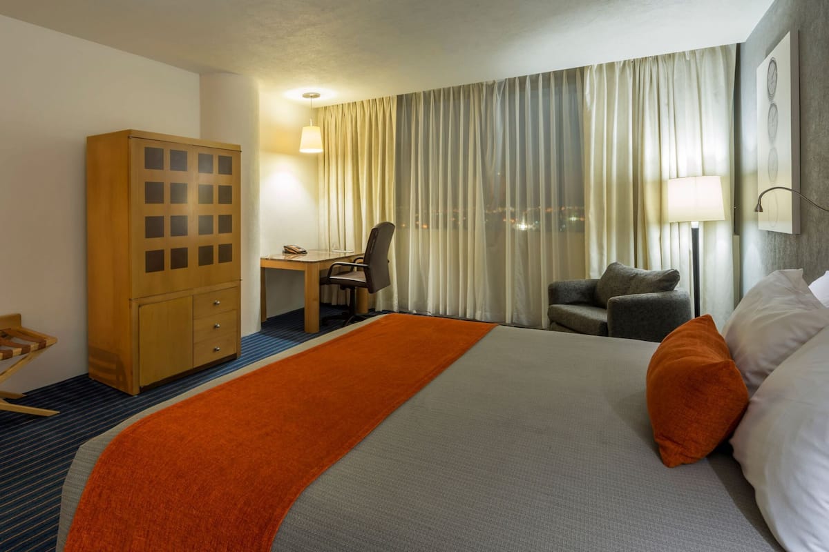 Una habitación de hotel con cama, escritorio, silla, armario y ventana con cortinas.