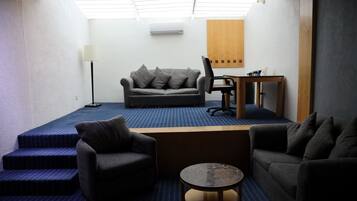 A compact living space with a sofa, two armchairs, a small table, and a desk. There is a lamp, a wooden panel on the wall, and a ceiling light.