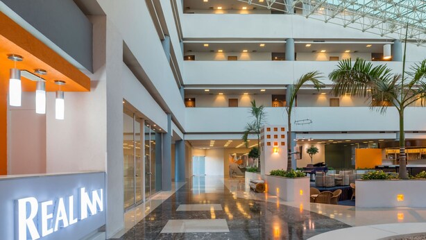 A multi-level hotel lobby with a glass ceiling, modern lighting, and a 'REAL IN' sign.